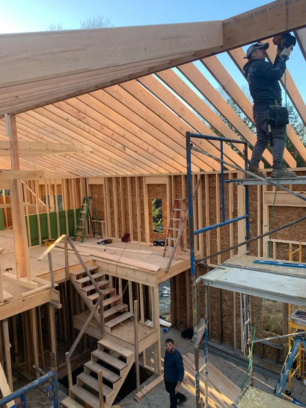 Construction workers framing a wooden roof structure with scaffolding and stairs.